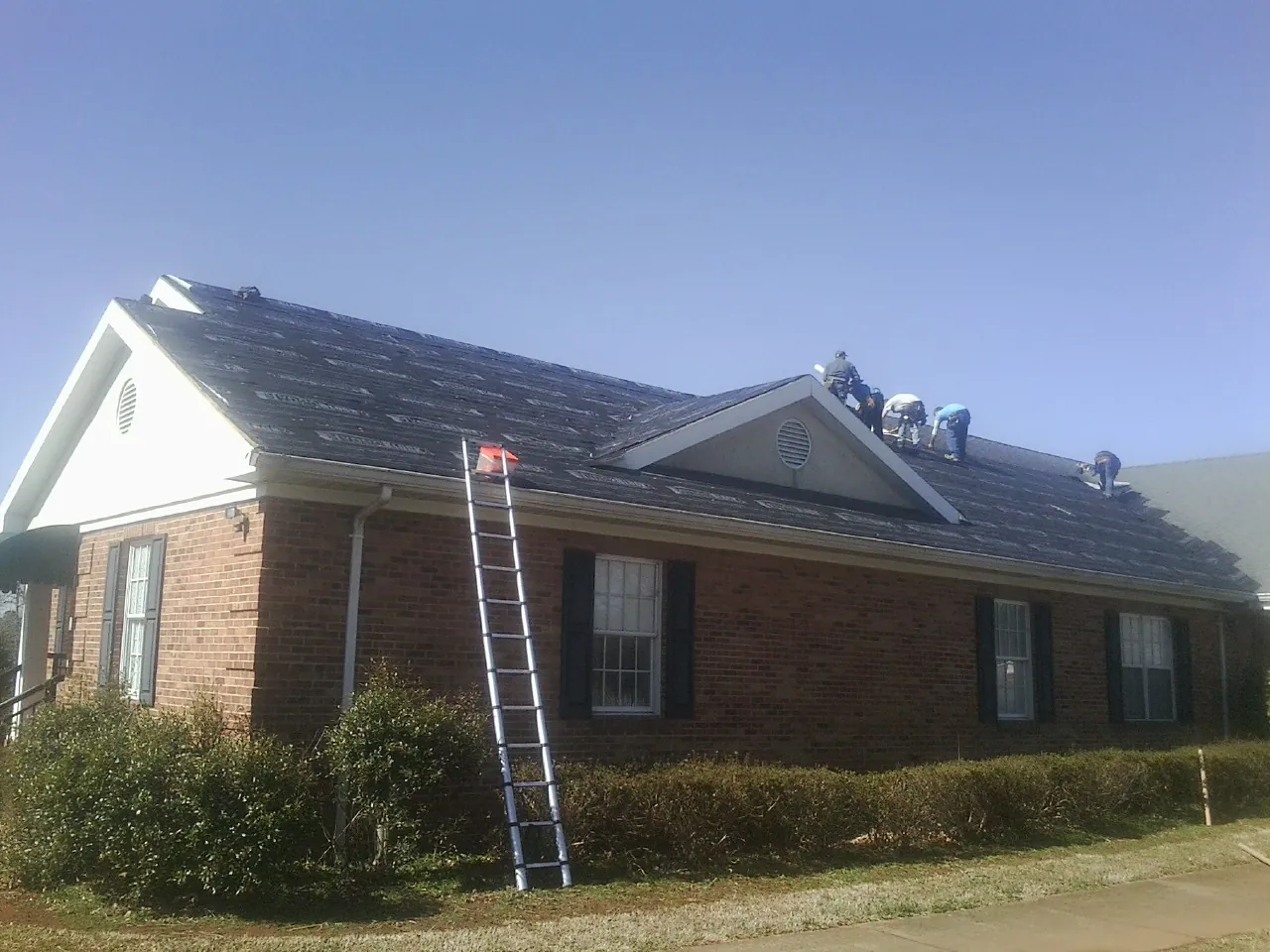 Crew tearing shingles off a brick ranch roof with a ladder set — Emergency Roof Repair work by our crew in Cheraw